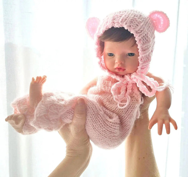 Bebé reborn silicona Iris sostenida en el aire con su gorrito de osita rosa, junto a la cortina