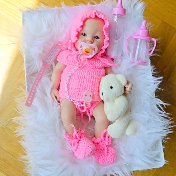 Bebé Reborn Silicona Sofia tumbada sobre alfombra blanca, con gorro y pelele rosa de Crochet, chupete, osito y biberones